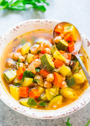 A bowl of hearty vegetable soup with zucchini, beans, carrots, and greens, served with a spoon.