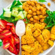 A bowl containing spiced chicken, roasted chickpeas, diced cucumber, halved cherry tomatoes, and a creamy sauce, with parsley garnish.
