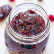 A jar of homemade cherry jam, with visible fruit pieces and seeds, against a white background with scattered cherries.