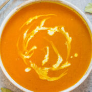 A bowl of creamy pumpkin soup with a swirl of cream, accompanied by a spoon and bay leaves on a textured surface.