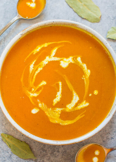 A bowl of creamy pumpkin soup with a swirl of cream, accompanied by a spoon and bay leaves on a textured surface.