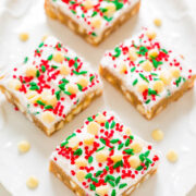 Four frosted sugar cookie bars with red, green, and white sprinkles and white chocolate chips on a white plate.
