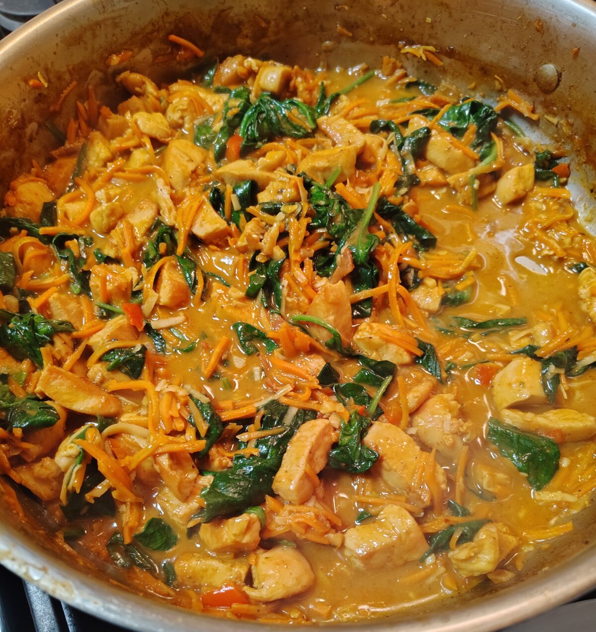 A pan containing a curry with chicken pieces, spinach, and shredded carrots in a creamy sauce.