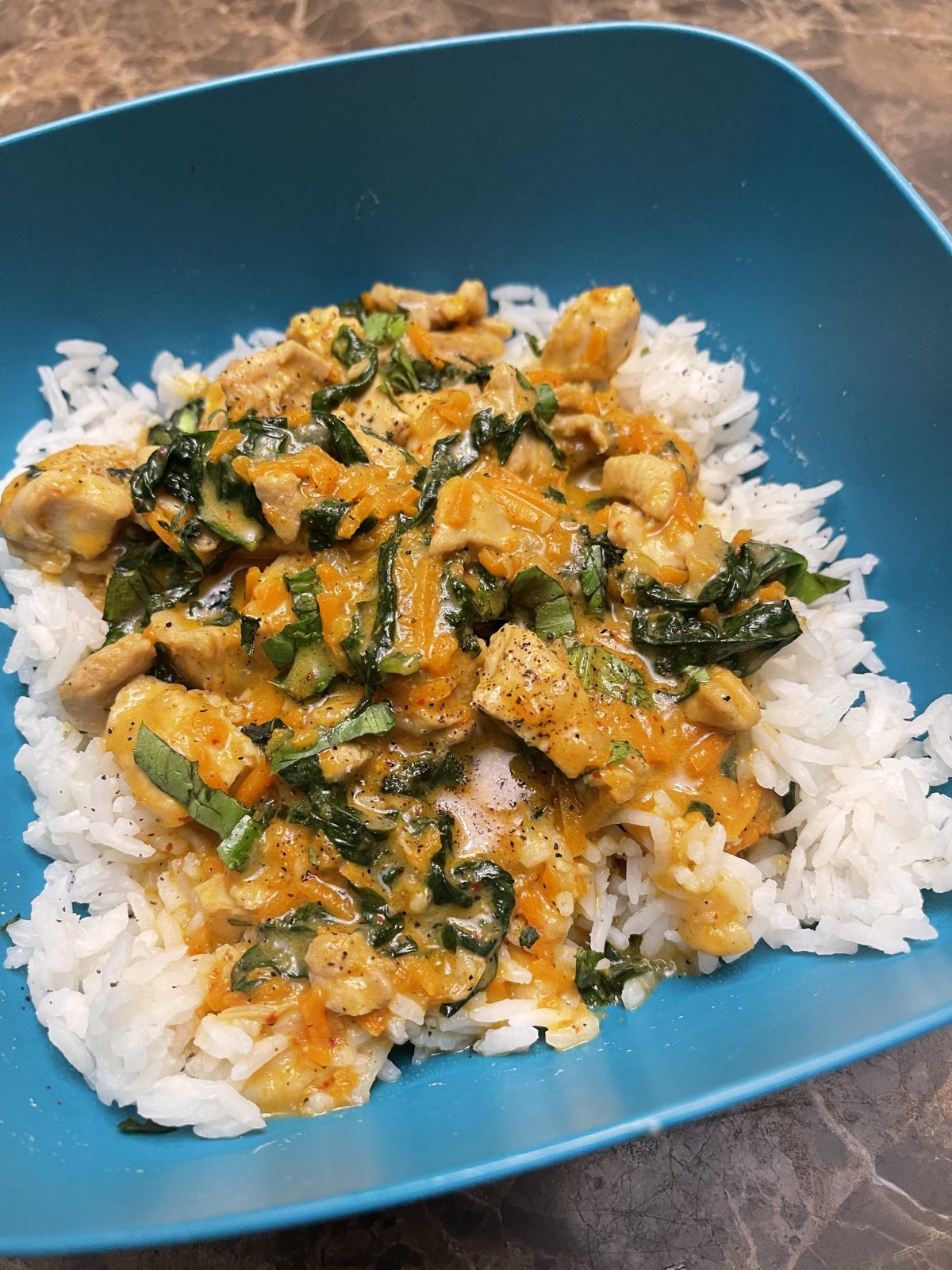 White rice topped with cooked chicken, spinach, and a creamy orange sauce, served in a blue bowl on a marble surface.