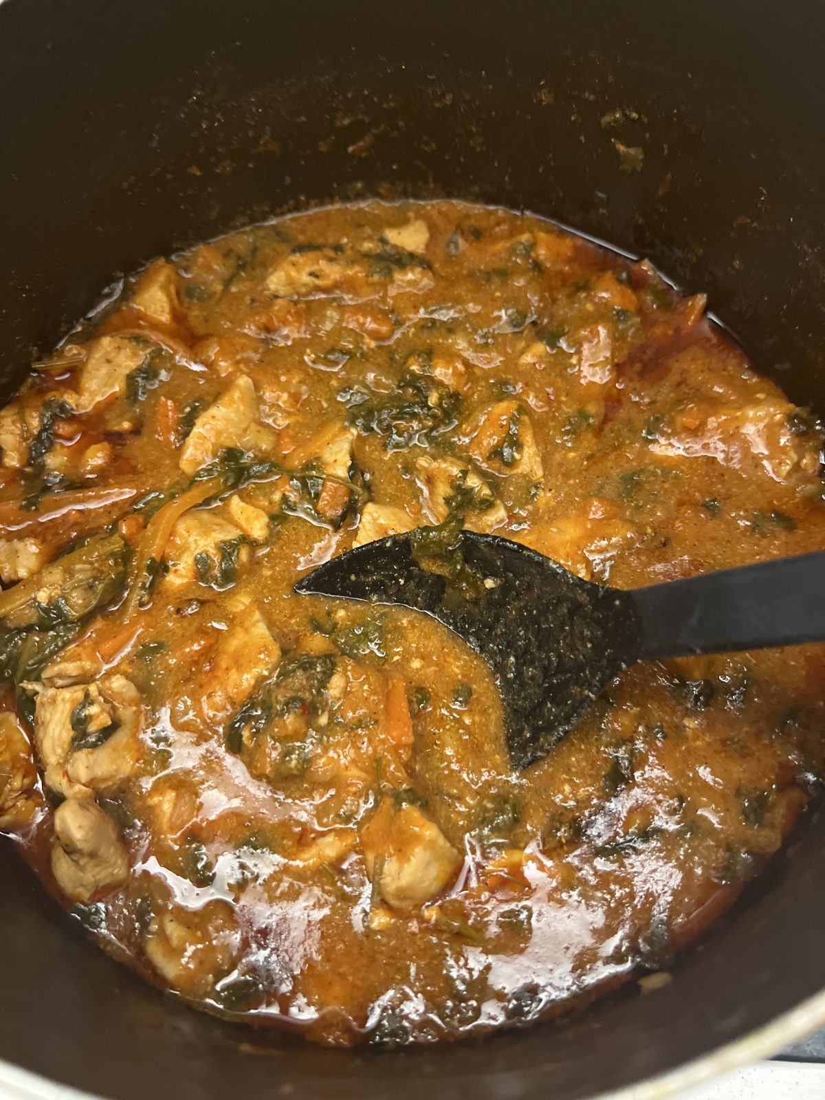 A pot of tomato-based stew with chunks of meat, green leafy vegetables, and a black spoon submerged in the mixture.