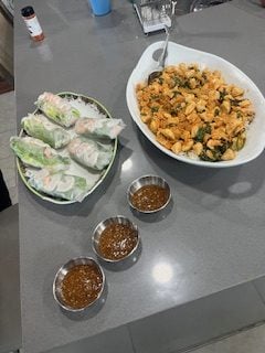 Spring rolls and a bowl of stir-fried noodles are placed on a kitchen counter, with three small bowls of dipping sauce arranged in front.