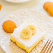 A slice of banana pudding dessert with vanilla wafers on a white ornate plate, accompanied by a fork on the side.