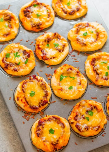 Mini cheese-topped pizzas on a baking sheet with some garnished with herbs.