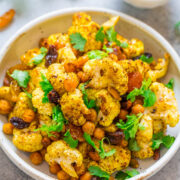 A bowl of roasted cauliflower and chickpeas garnished with herbs and served with a side of sauce.
