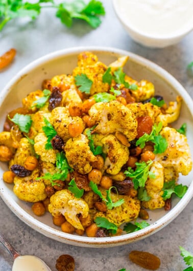 A bowl of roasted cauliflower and chickpeas garnished with herbs and served with a side of sauce.
