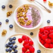 Bowls of yogurt topped with granola, chia seeds, and mixed berries on a white wooden surface.