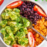 A colorful bowl of food containing pesto-coated chicken, black beans, rice, and sliced tomatoes, garnished with fresh cilantro.