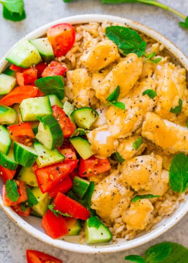 A bowl of brown rice topped with diced chicken, cucumber, and tomato garnished with fresh herbs.