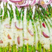 Baked asparagus with melted cheese and red onion slices on a baking sheet.