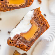 A chocolate brownie with caramel filling and cream topping, placed on a white surface with chocolate sprinkles and additional caramel pieces nearby.