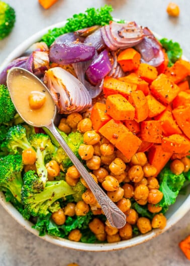 A bowl filled with roasted vegetables and chickpeas, with a spoon drizzling sauce over the top.