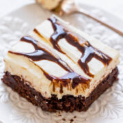 A slice of layered dessert with brownie base and cream topping drizzled with chocolate syrup on a white plate.