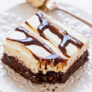 A slice of layered dessert with brownie base and cream topping drizzled with chocolate syrup on a white plate.