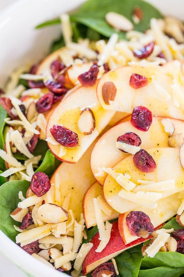 A salad with fresh spinach, sliced apples, shredded cheese, dried cranberries, and sliced almonds.