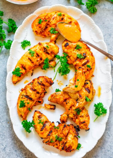 Grilled chicken tenderloins with a side of sauce, garnished with parsley on a white plate.