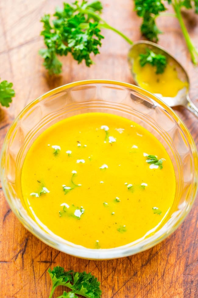 A clear glass bowl of yellow soup garnished with crumbled white cheese and chopped parsley sits on a wooden surface with a spoon and parsley sprigs nearby.