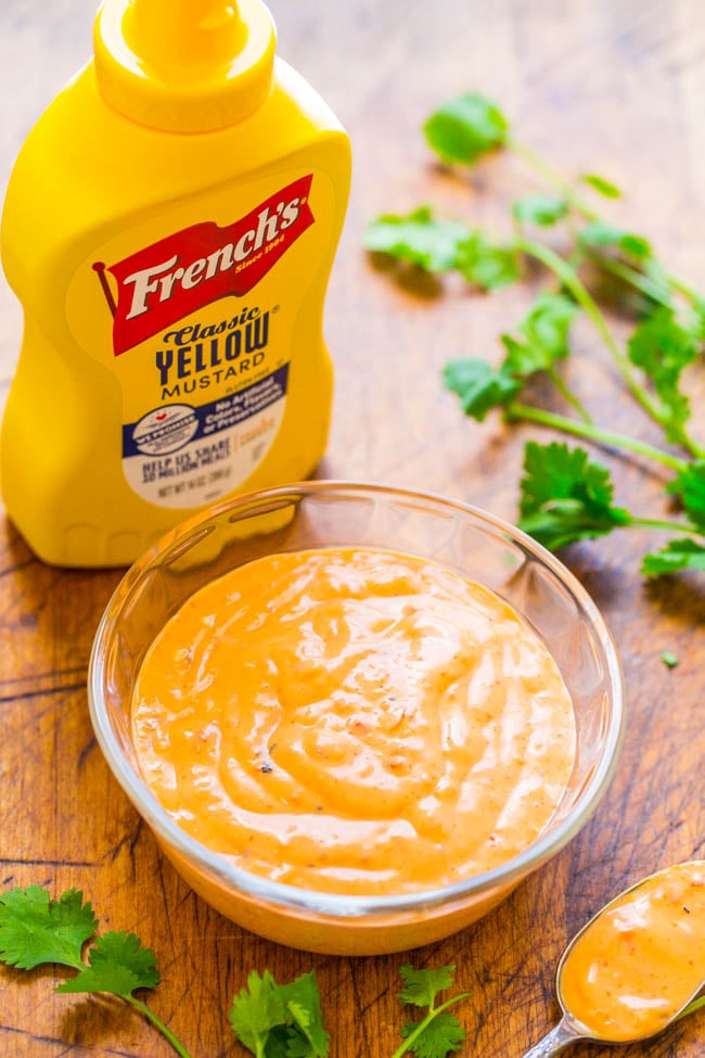 A bottle of Frenchโs Classic Yellow Mustard next to a glass bowl of creamy orange sauce, with cilantro leaves scattered on a wooden surface.
