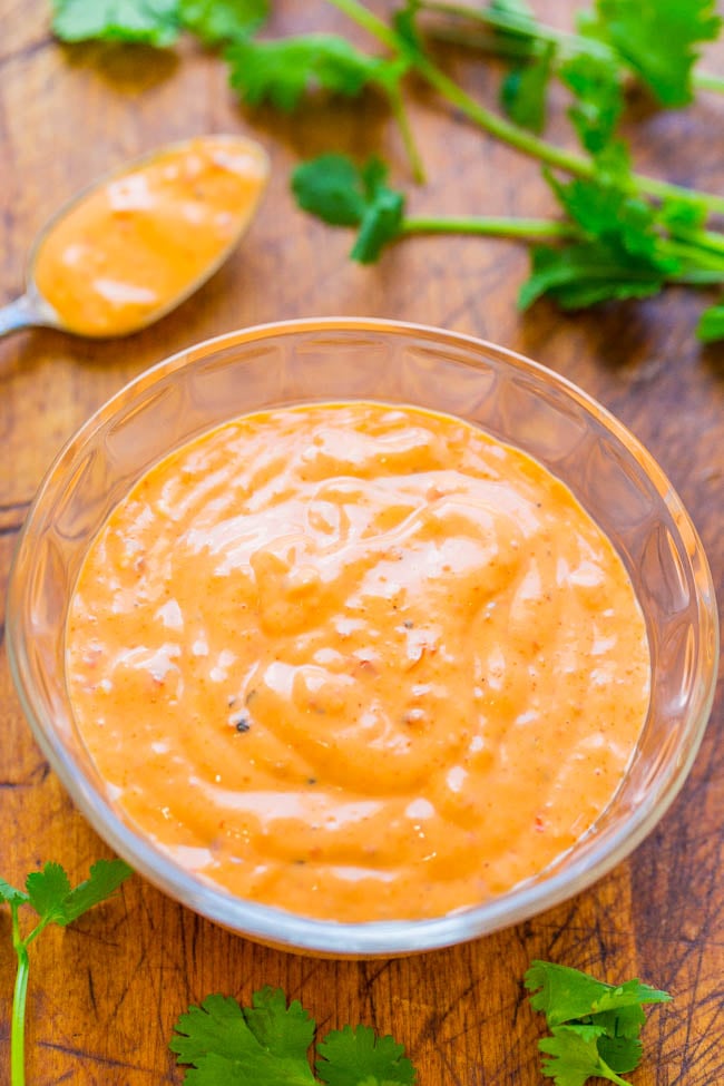 A glass bowl filled with creamy orange sauce sits on a wooden surface, with a spoonful of sauce and fresh cilantro leaves nearby.
