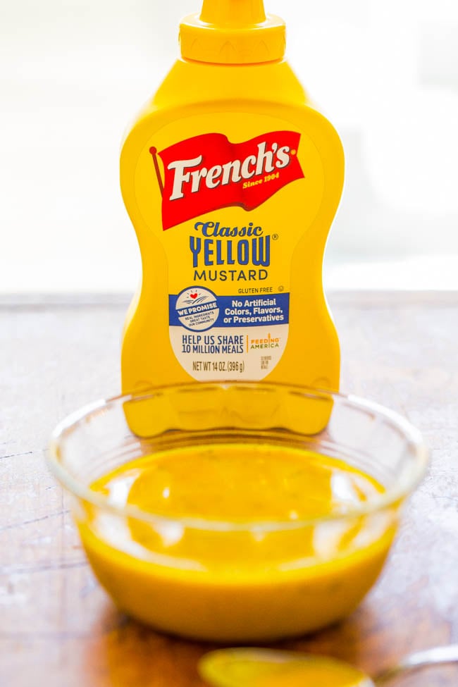 A bottle of French's Classic Yellow Mustard stands behind a glass bowl filled with mustard on a wooden surface.