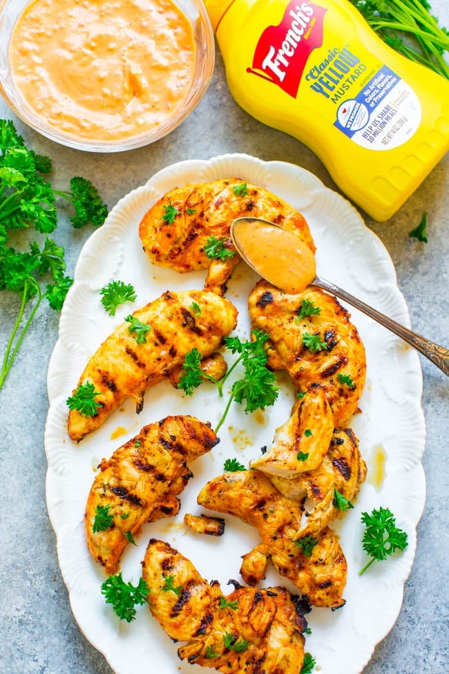 Grilled chicken tenders on a white plate, garnished with parsley, served with a spoonful of sauce. Frenchโs yellow mustard and a bowl of sauce are visible nearby.