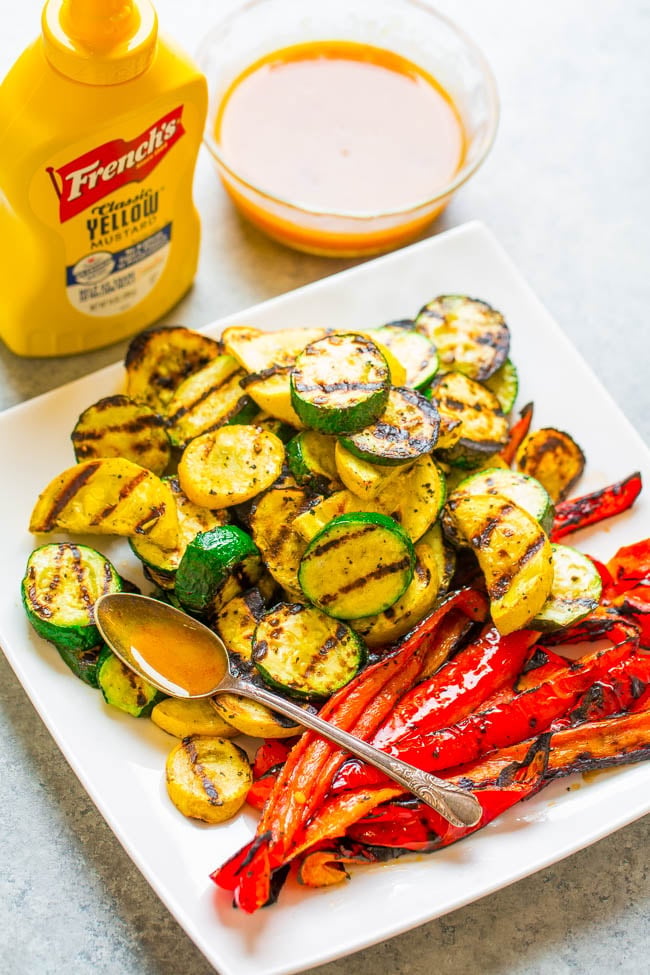 A plate of grilled zucchini and red bell peppers with a spoon, served alongside a bowl of sauce and a bottle of Frenchโs yellow mustard.