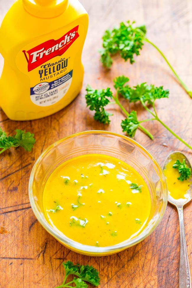 A bowl of yellow mustard sauce garnished with herbs sits on a wooden surface beside a French's mustard bottle, parsley sprigs, and a spoon.