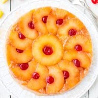 A pineapple upside-down cake garnished with cherries, displayed on a white plate with forks on the side.