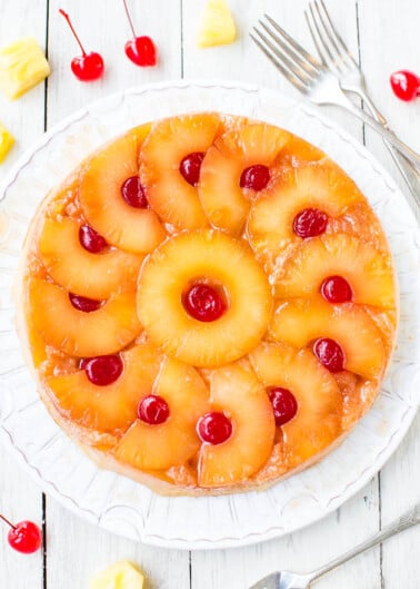 A pineapple upside-down cake garnished with cherries, displayed on a white plate with forks on the side.