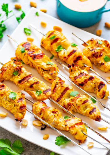 Grilled chicken skewers garnished with cilantro and peanuts on a white platter.