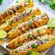 Grilled corn on the cob dressed with creamy sauce, spices, cheese, and cilantro, garnished with lime wedges on the side.