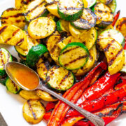 Grilled zucchini and red bell peppers on a white plate with a spoon drizzling dressing.
