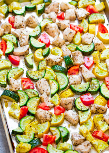Seasoned chicken pieces and sliced zucchini and tomatoes arranged on a baking sheet.