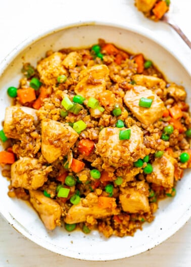 A bowl of stir-fried rice with chicken, carrots, peas, and garnished with green onions.