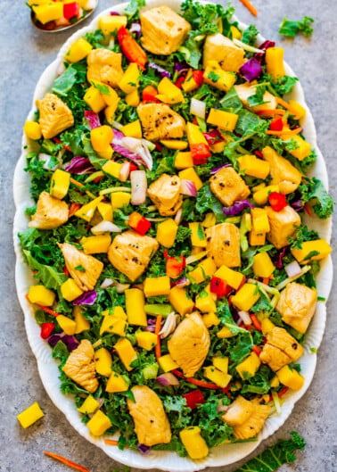 A colorful grilled chicken salad with kale, mango, and bell peppers served on a white platter.