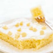 A slice of lemon bar on a white plate garnished with powdered sugar and small pieces of lemon peel.