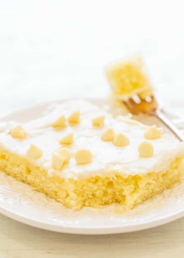 A slice of lemon bar on a white plate garnished with powdered sugar and small pieces of lemon peel.