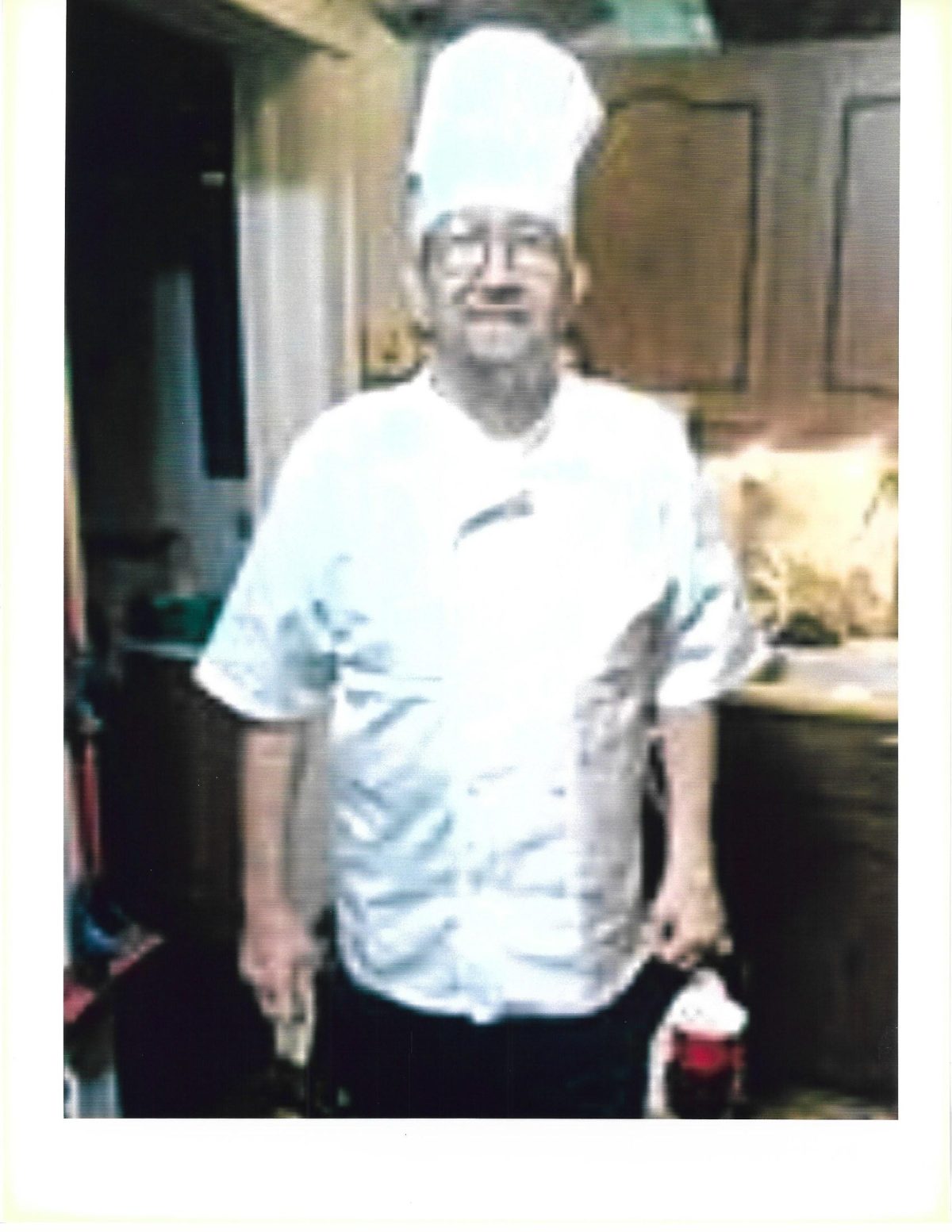 A man wearing a white chef’s coat and hat stands in a kitchen, holding items in both hands, facing the camera.