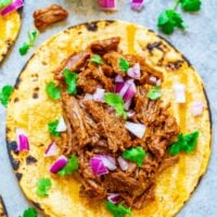 A corn tortilla topped with shredded beef, diced red onions, and fresh cilantro, served with a lime wedge.