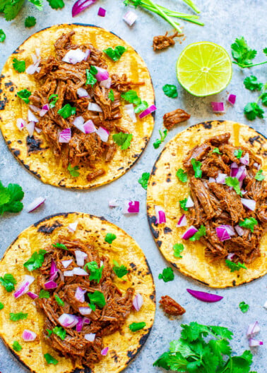 Three beef tacos garnished with red onion and cilantro on a speckled surface, with lime wedges and herbs scattered around.