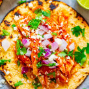 A tostada topped with shredded chicken, diced onions, cilantro, cheese, and hot sauce, garnished with a lime wedge on the side.
