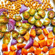 Roasted vegetables including chickpeas, brussels sprouts, red onions, and sweet potatoes on a baking sheet.