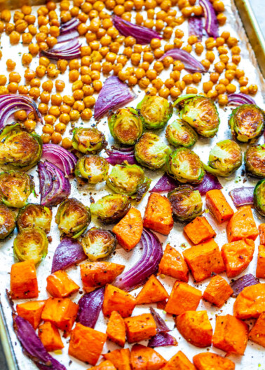 Roasted vegetables including chickpeas, brussels sprouts, red onions, and sweet potatoes on a baking sheet.