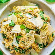 A bowl of pasta with spinach, artichokes, and shaved parmesan cheese on a white surface.
