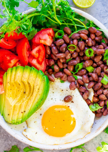 A bowl containing a healthy meal with sliced avocado, beans, tomato, herbs, a sunny-side-up egg, and a lime wedge.