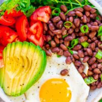 A colorful bowl of food with avocado, beans, tomatoes, a fried egg, and fresh herbs.